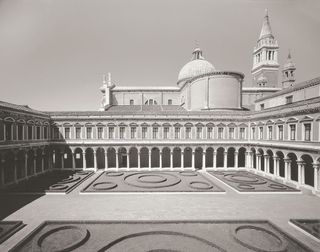 Black and white image of the Church of San Giorgio Maggiore, Venice, tall dome roof tops with bell towers, courtyard with row of stone arches a long the edge of the centre piece of circle stone designs inside rectangle borders, windows above the arches, clear sky
