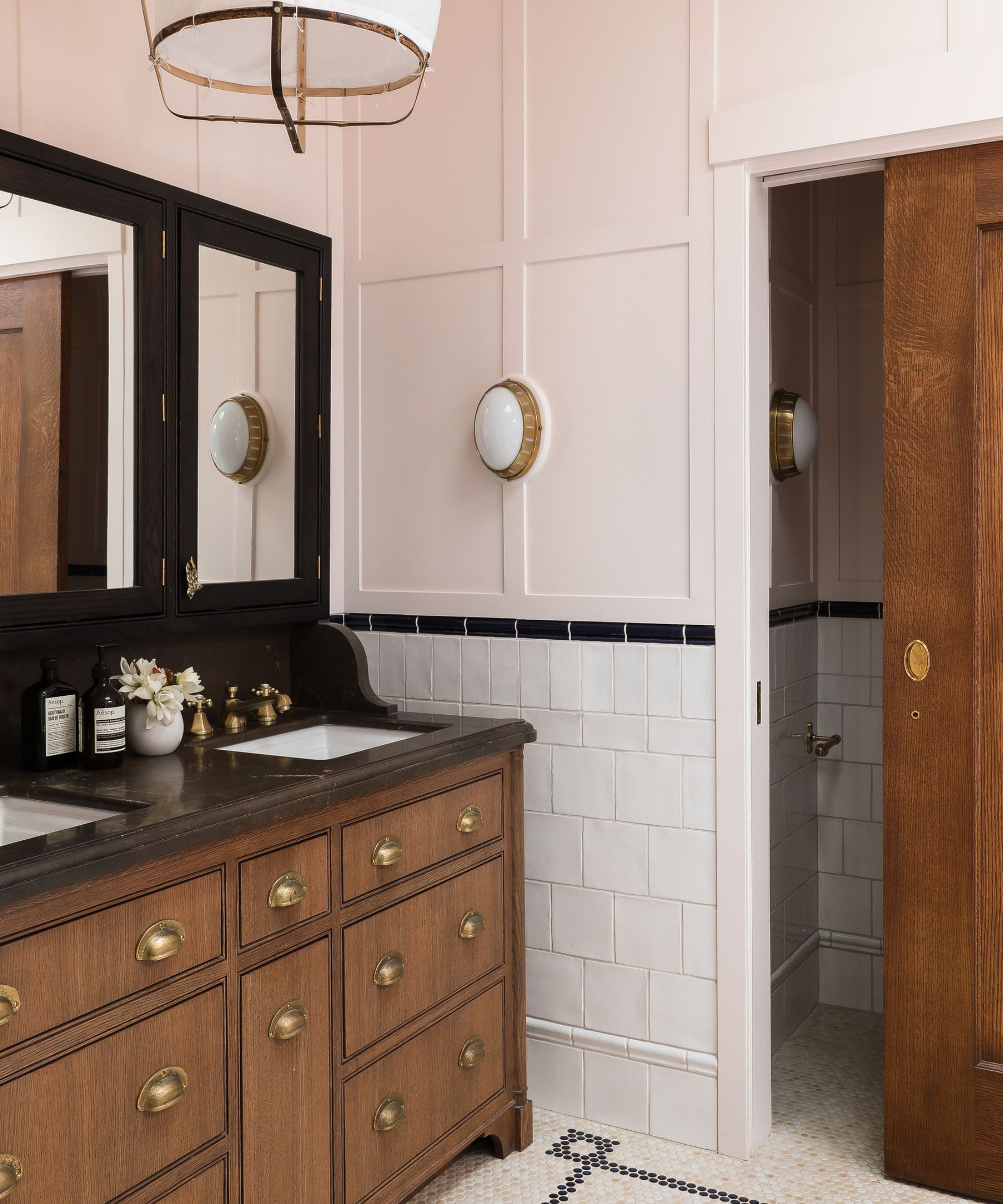 A bathroom with peach pink panelled walls, white tiles on the lower walls, a large wooden vanity with dark brown countertops, double sinks and mirrors, and a wooden sliding door.