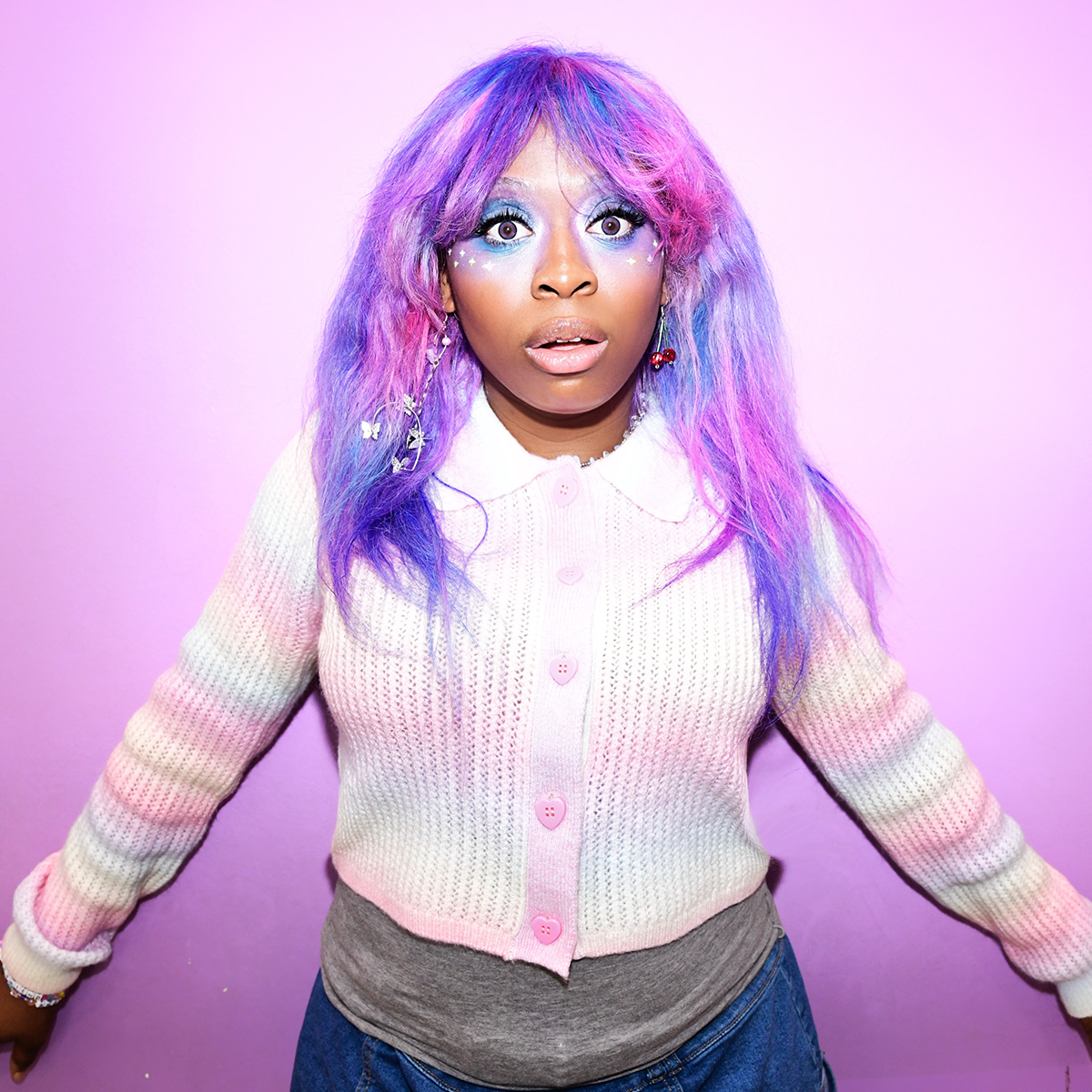 Artist hemlocke springs is photographed against a purple backdrop. She is wearing a rainbow-colored cardigan with pink, heart-shaped buttons, and her hair is bright pink and purple.