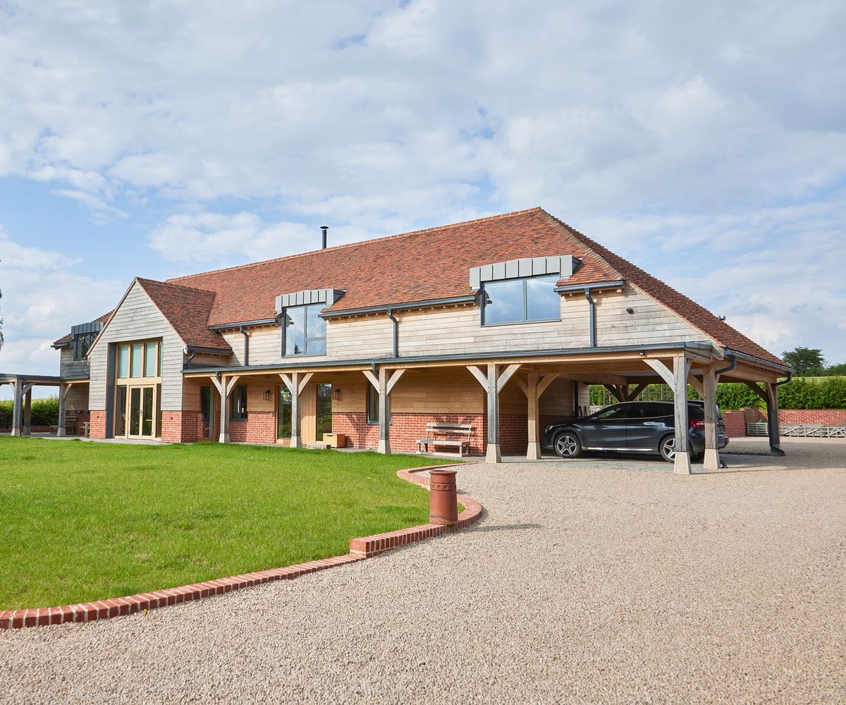 A large oak frame home with a gravel driveway leading to a covered parking area for cars. There is a lawn to the left