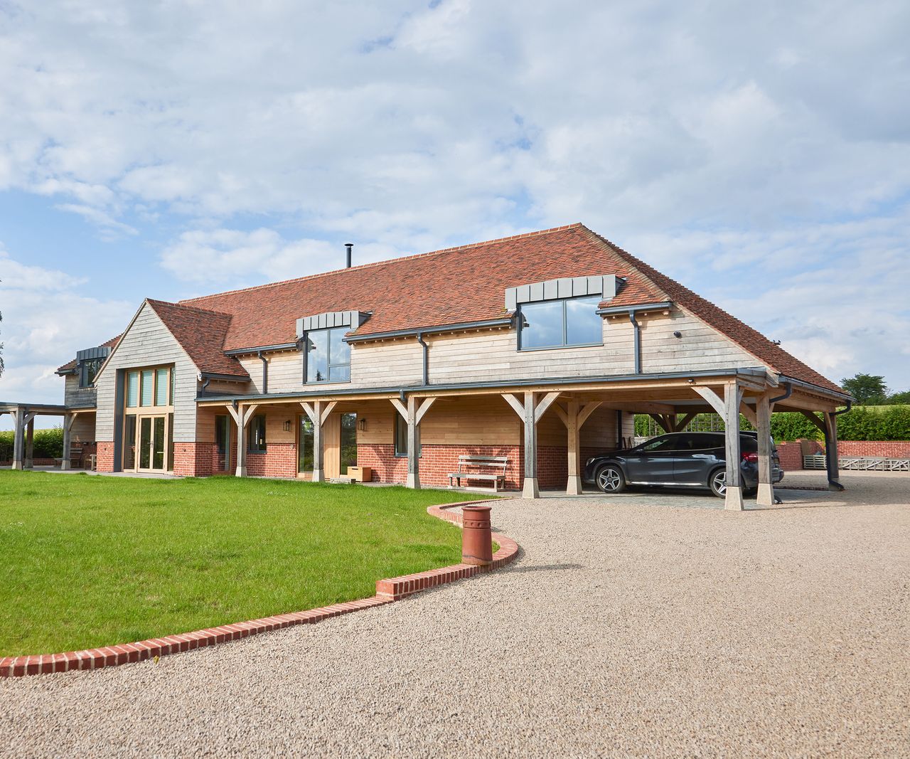 A large oak frame home with a gravel driveway leading to a covered parking area for cars. There is a lawn to the left