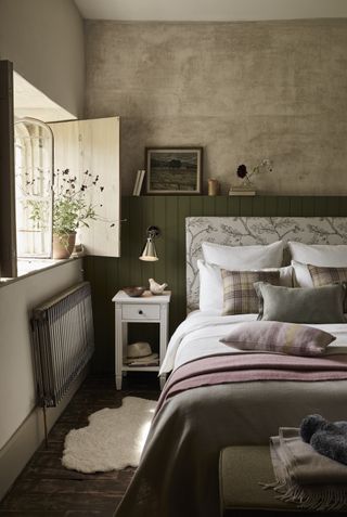 bedroom with neutral textured walls, green panelling behind bed, upholstered fabric headboard, tartan cushions, cottage style, wooden floor, bedside table, wall light