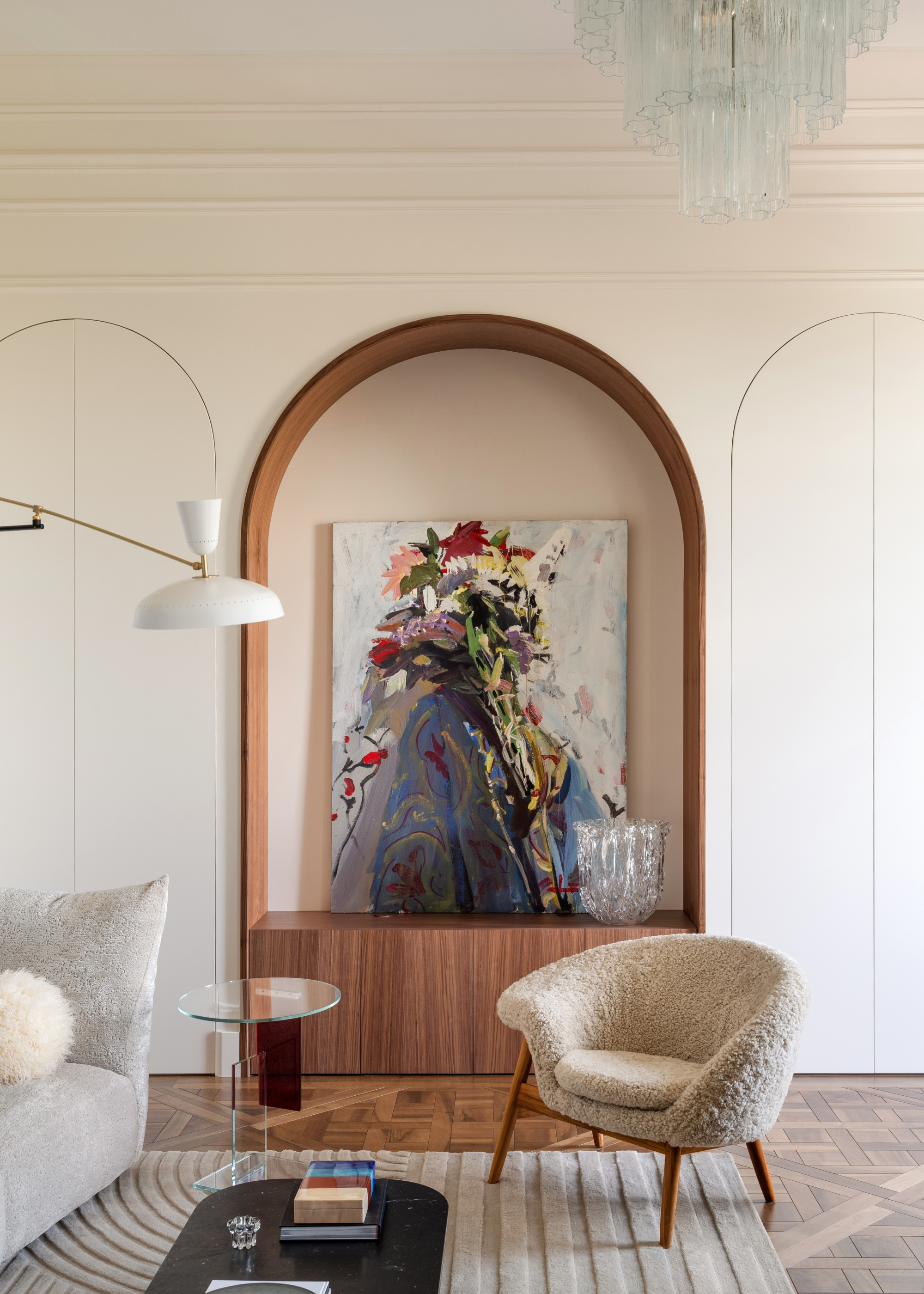 a modern living area with three arches on the back wall, one in the center which is highlighted with wood on the base and inside the arch, inside of which is a colorful piece of art, the other two arches are closed, hidden doors with an accent chair and table in front
