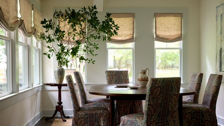 A light-filled sunroom with a round wooden dining table, upholstered dining chairs, linen Roman blinds, a large leafy arrangement in a ceramic vase and wraparound windows overlooking the garden.