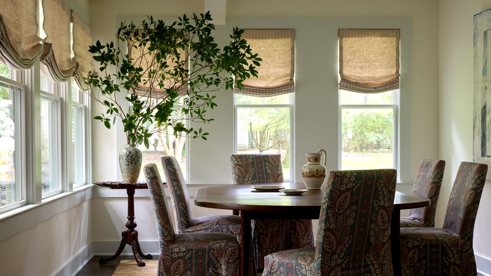 A light-filled sunroom with a round wooden dining table, upholstered dining chairs, linen Roman blinds, a large leafy arrangement in a ceramic vase and wraparound windows overlooking the garden.