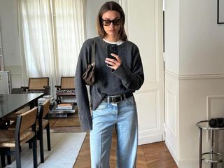 French fashion influencer Anne-Laure Mais poses for a mirror selfie in her chic Paris apartment wearing black sunglasses, a charcoal gray crewneck sweater layered over a white T-shirt, a mini Louis Vuitton chain shoulder bag, a black belt, and light-wash jeans.