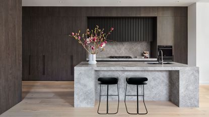 A dark wood kitchen with light wood floor and concrete effect island with black bar stools