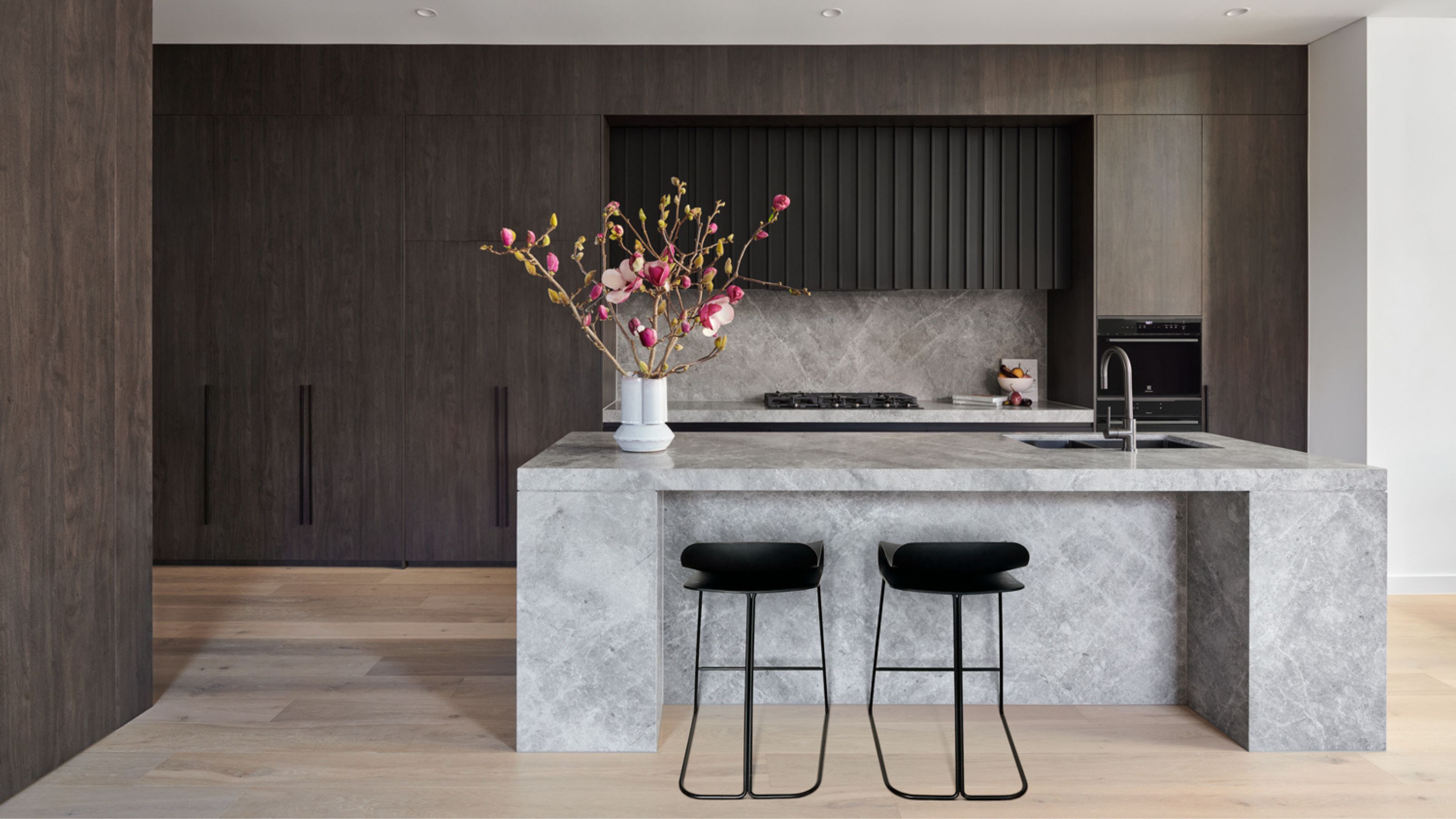A dark wood kitchen with light wood floor and concrete effect island with black bar stools