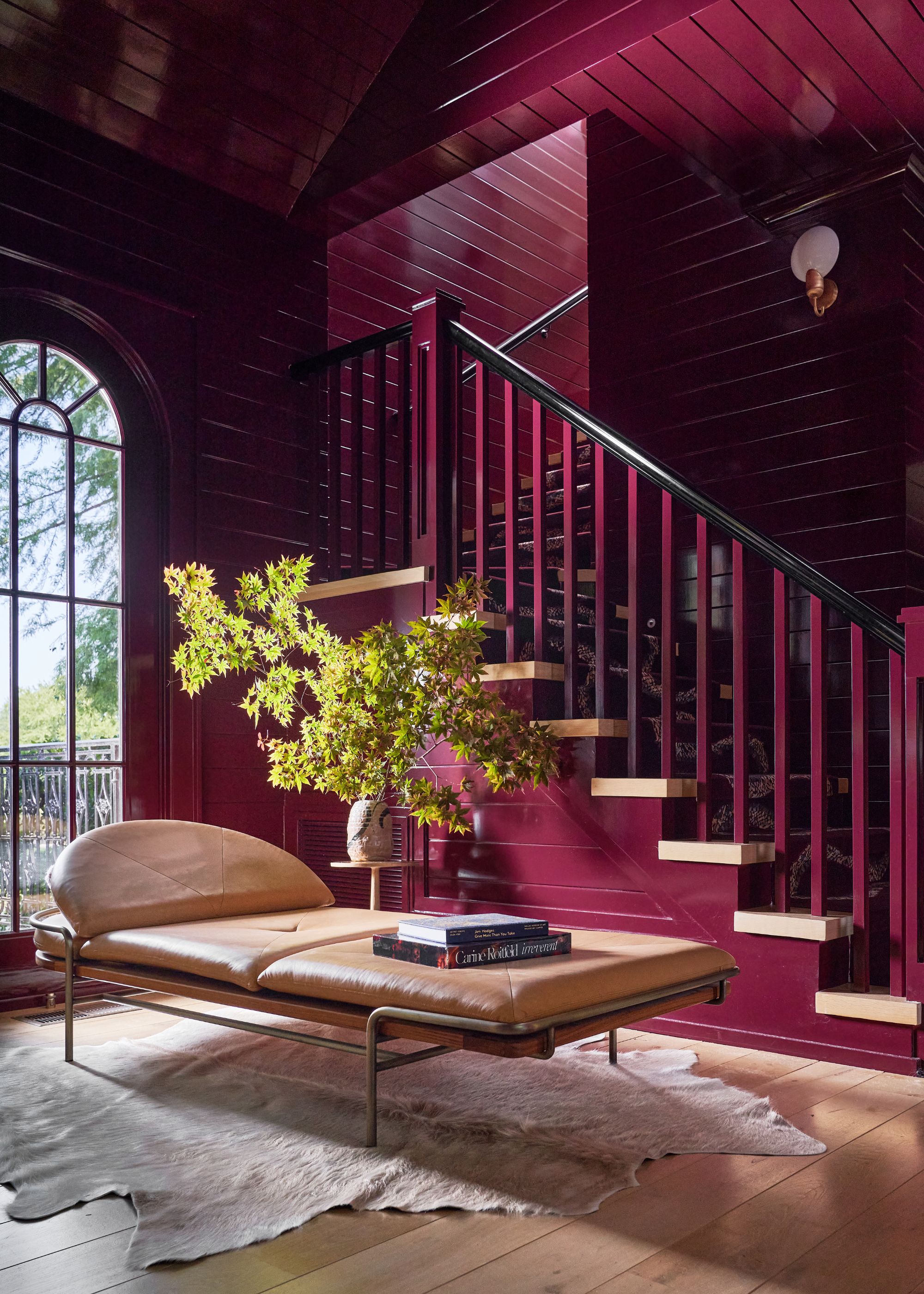A living space with a staircase and tall arched window, color-drenched in a rich shade of burgundy, wood flooring with a rug, a modern leather seat, and a large vase of green foliage.