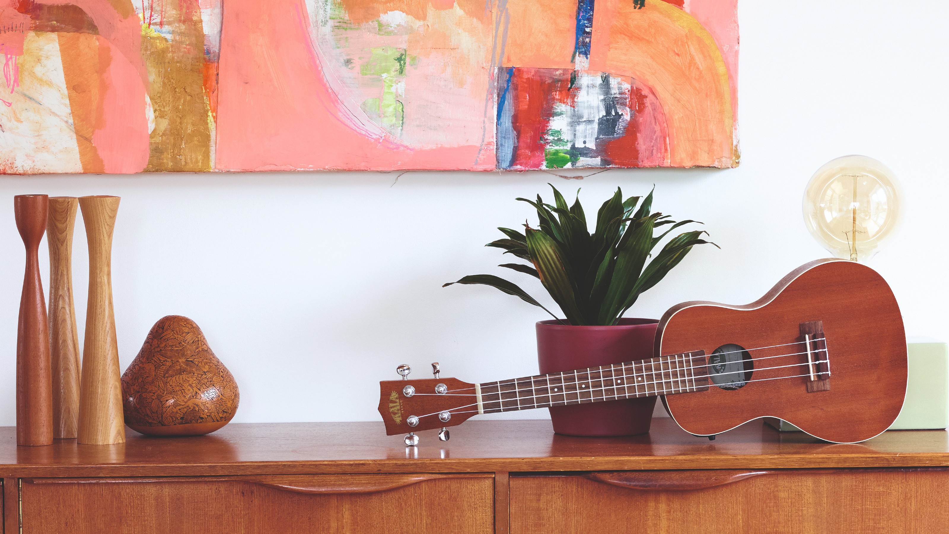 A midcentury sideboard with an abstract artwork hung above, styled with a set of wooden vases, a plant, a guitar and a table lamp