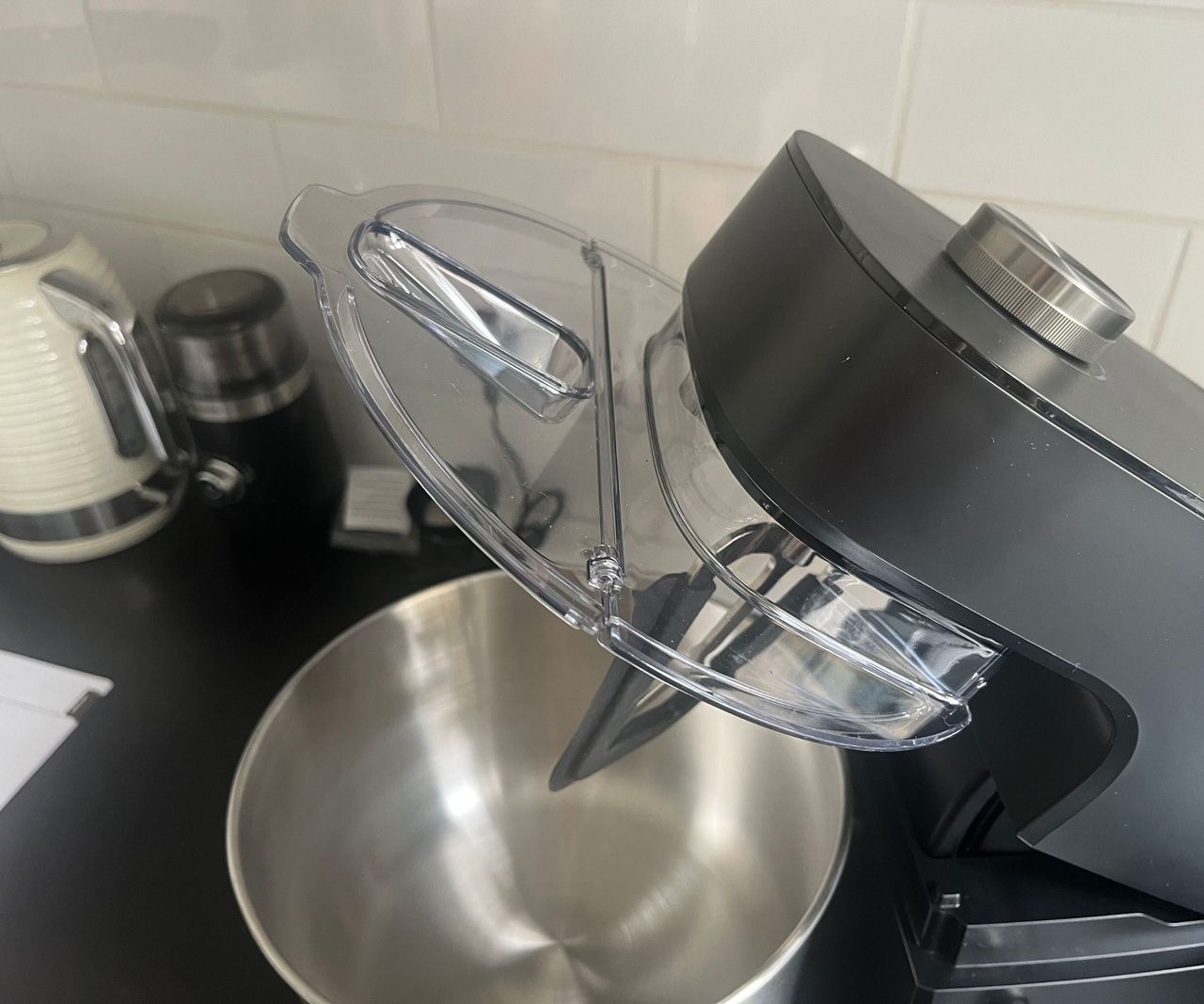 A close up of a clear plastic spill lid attached to the tilt head of a black stand mixer
