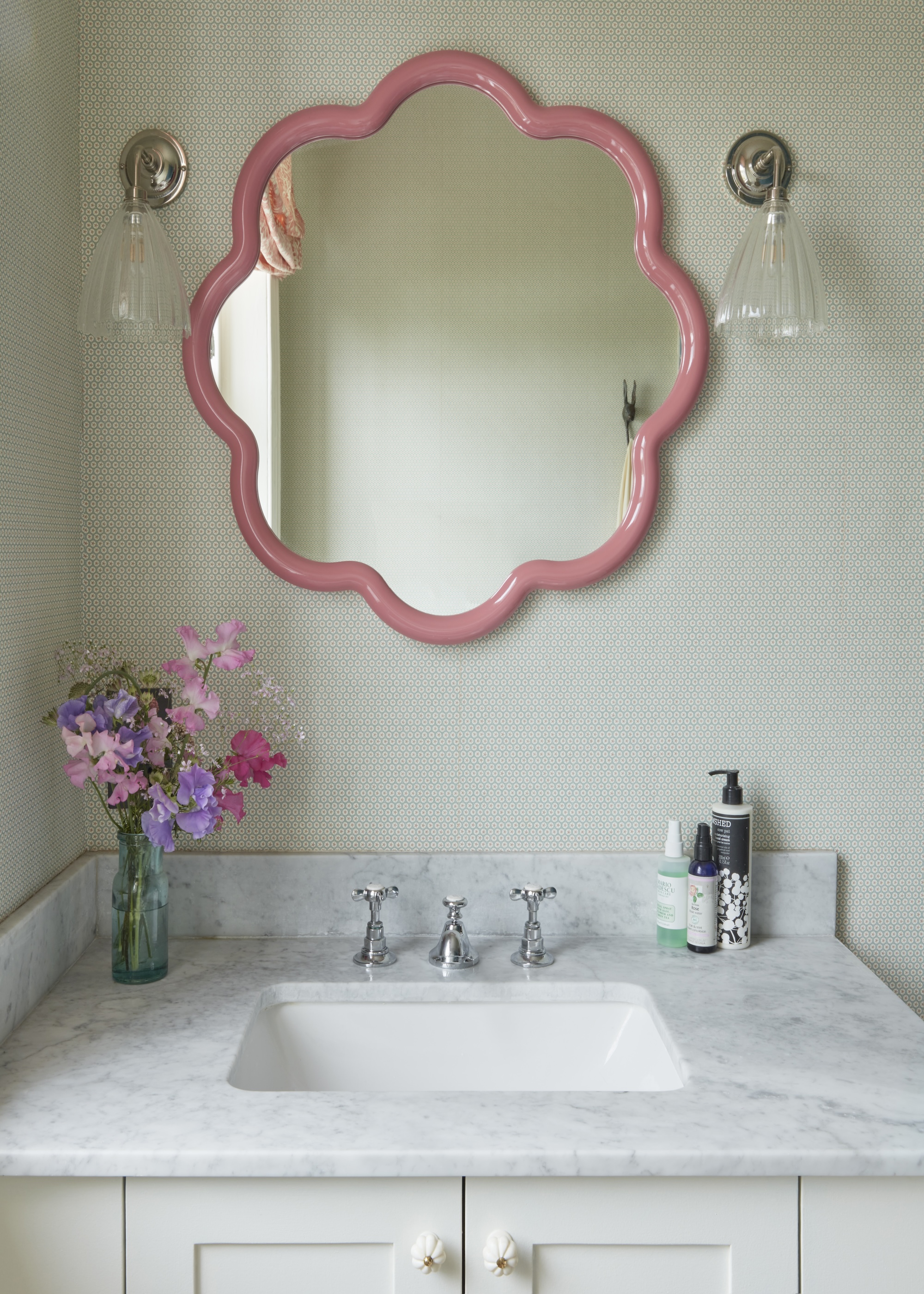 A pink curvy mirror with a small white marble sink with toiletries and a vase with flowers inside.