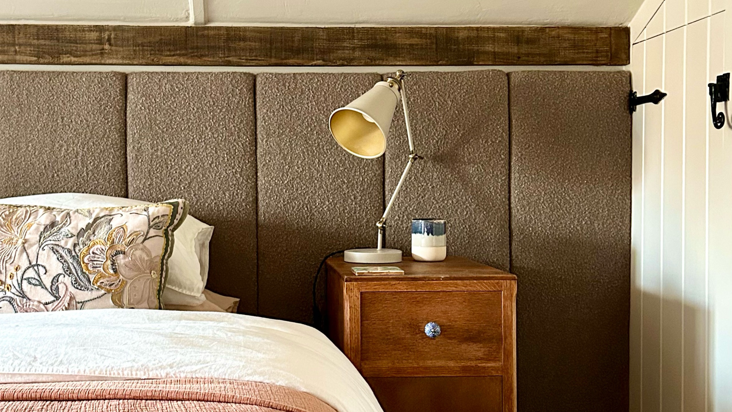 Country eaves bedroom with wide upholstered panelled headboard, wooden bedside table, lamp and pink throw