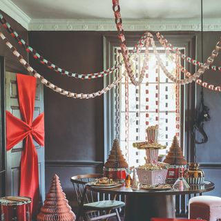 A dining room with a round table decorated for Christmas with the ceiling and window draped with patterned paper chains