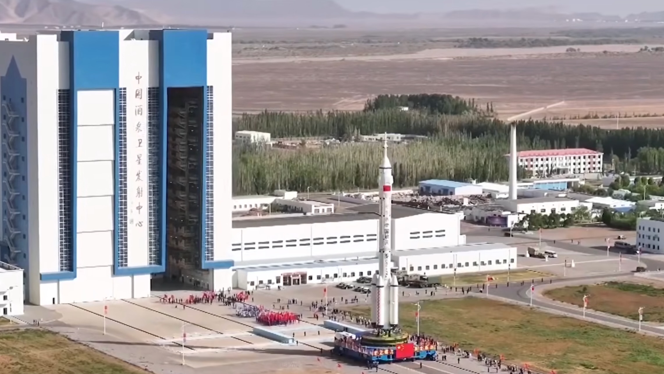 long-distance aerial photo of a large white rocket emerging from a big white building onto an outdoor tarmac, with lots of people around