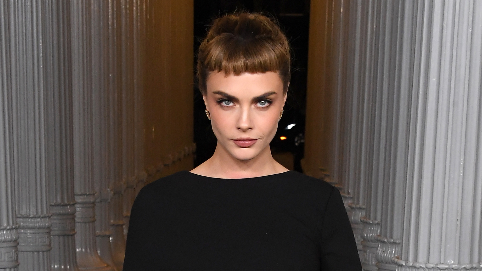 Cara Delevigne with an updo hairstyle that has bangs. She's also wearing a black dress.