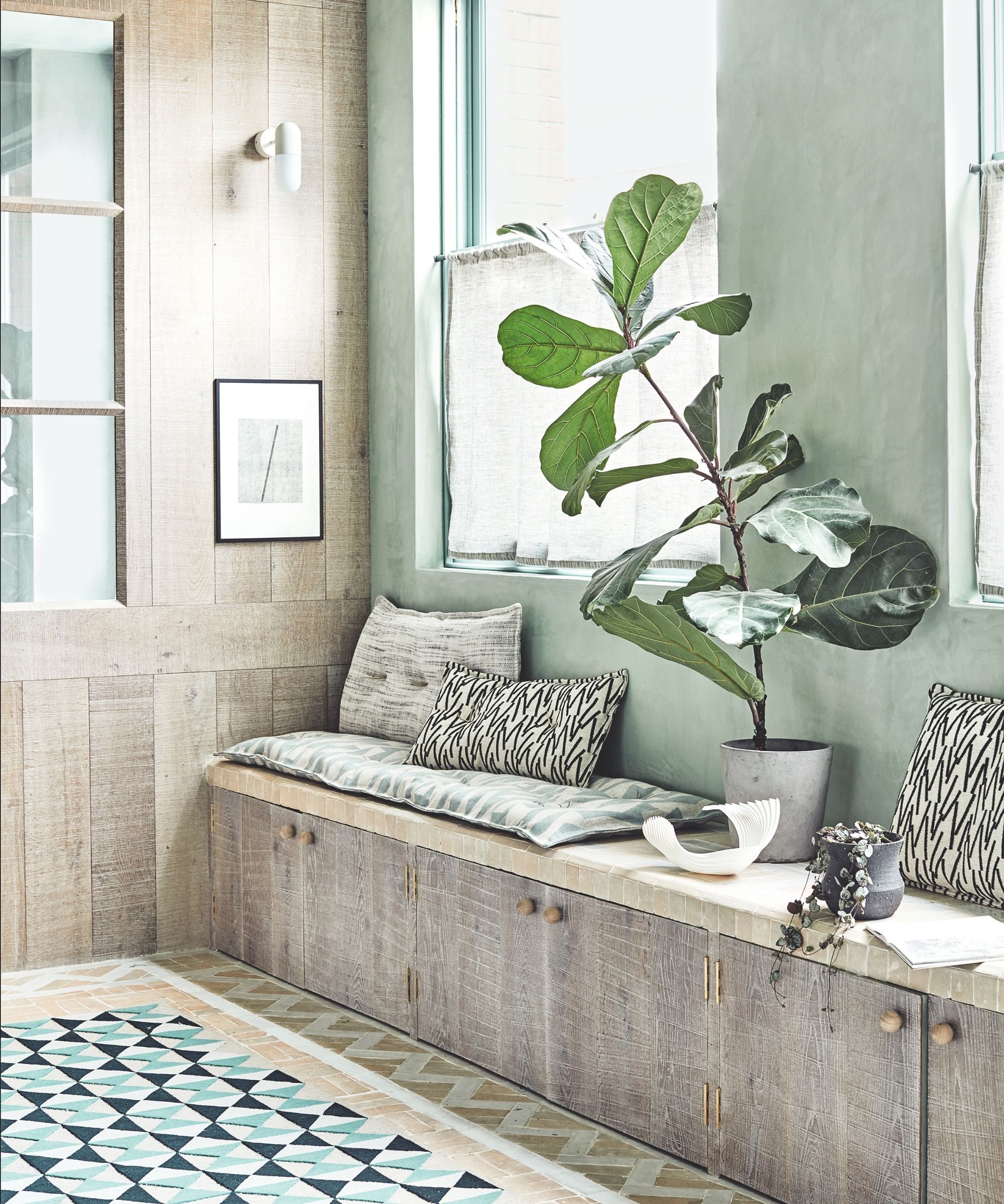 A wooden and sage green seating area with large plant, tiled flooring, patterned cushions, and large windows.