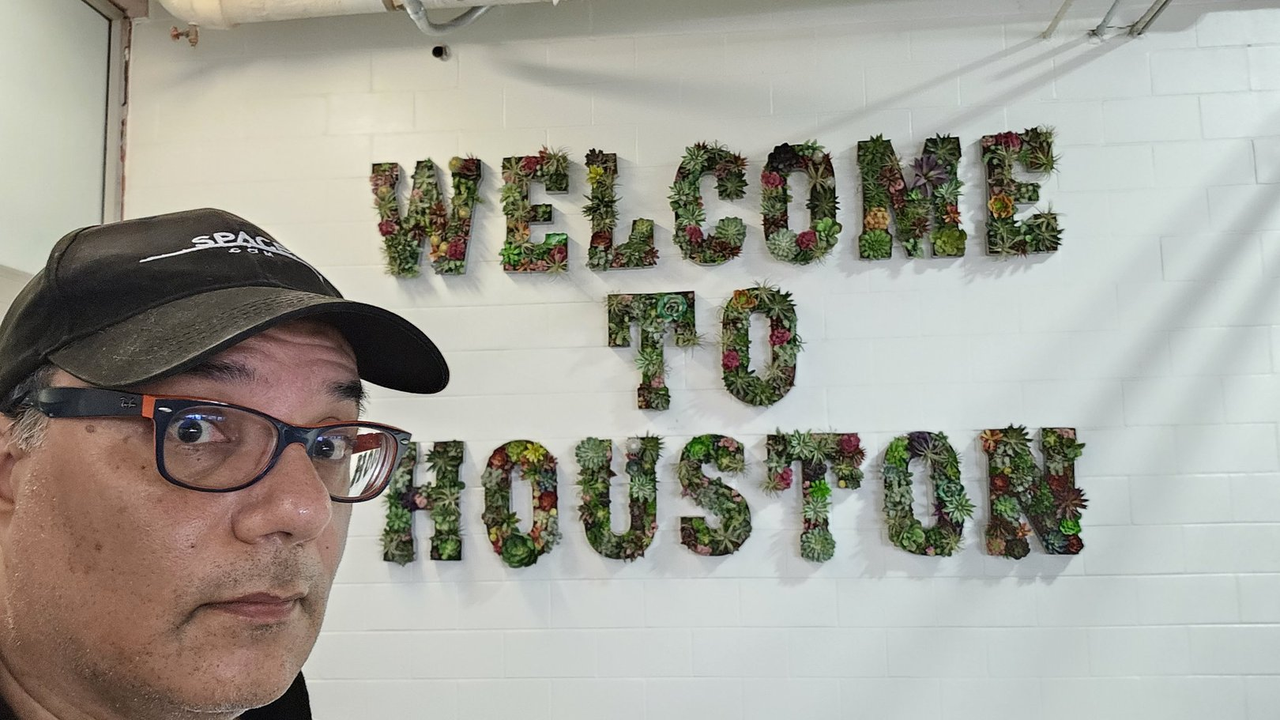 A man looks at the camera while standing in front of a sign that reads "welcome to Houston" on a white wall.