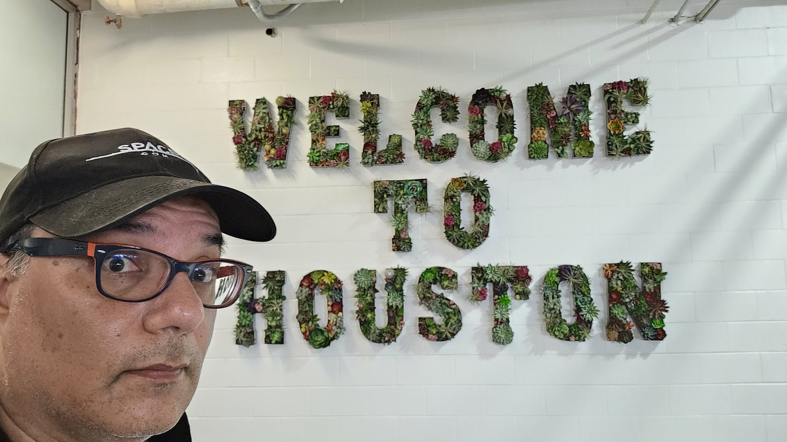 A man looks at the camera while standing in front of a sign that reads "welcome to Houston" on a white wall.