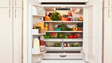 An open fridge with fruit and vegetables on the shelves and milk and juice bottles in the door, with cream cabinet doors around it