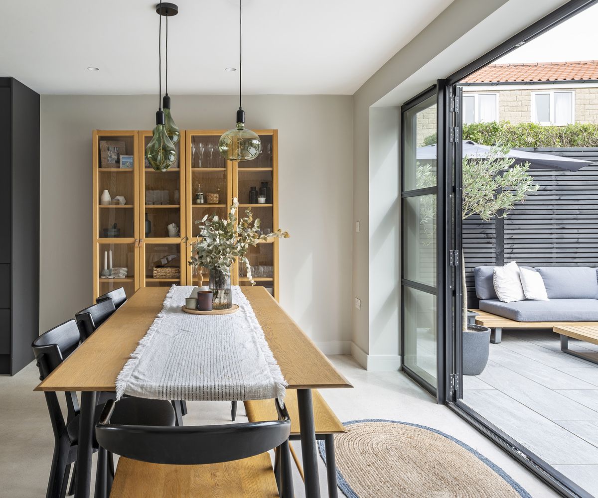 A beautifully styled dining table which looks out through open glass patio doors to a garden. The interior style is Japandi with pale wood and flooring with dark accents