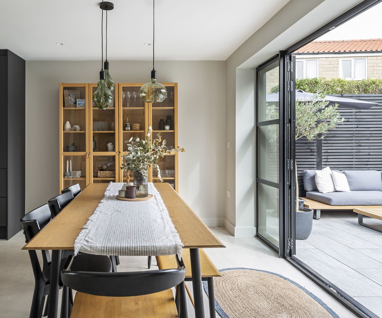A beautifully styled dining table which looks out through open glass patio doors to a garden. The interior style is Japandi with pale wood and flooring with dark accents