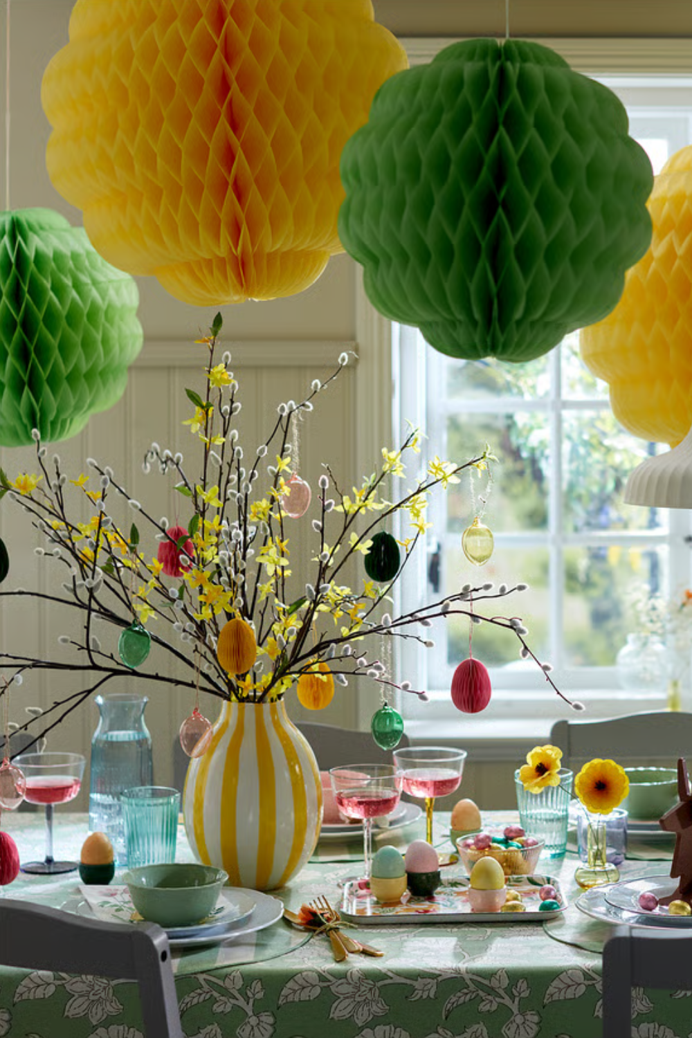 Cheerful Easter tablescape featuring a yellow floral centerpiece adorned with pastel egg ornaments, multi-colored stemware, scalloped ceramics, and a cluster of honeycomb paper decorations hanging overhead.