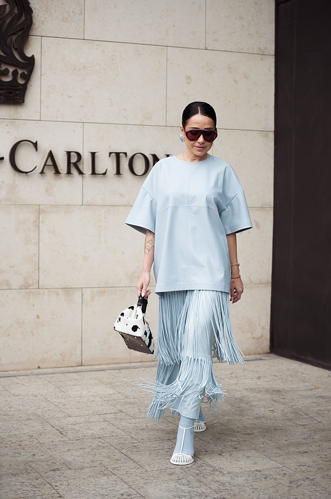 Elena Kolisnichenko, Visa Fashion Week Almaty 2025'in 3. gününde püsküllü açık mavi uzun maksi etek, mavi deri üst, mavi tayt, beyaz ayakkabılar, inek desenli çanta ve kahverengi güneş gözlüğü giyiyor
