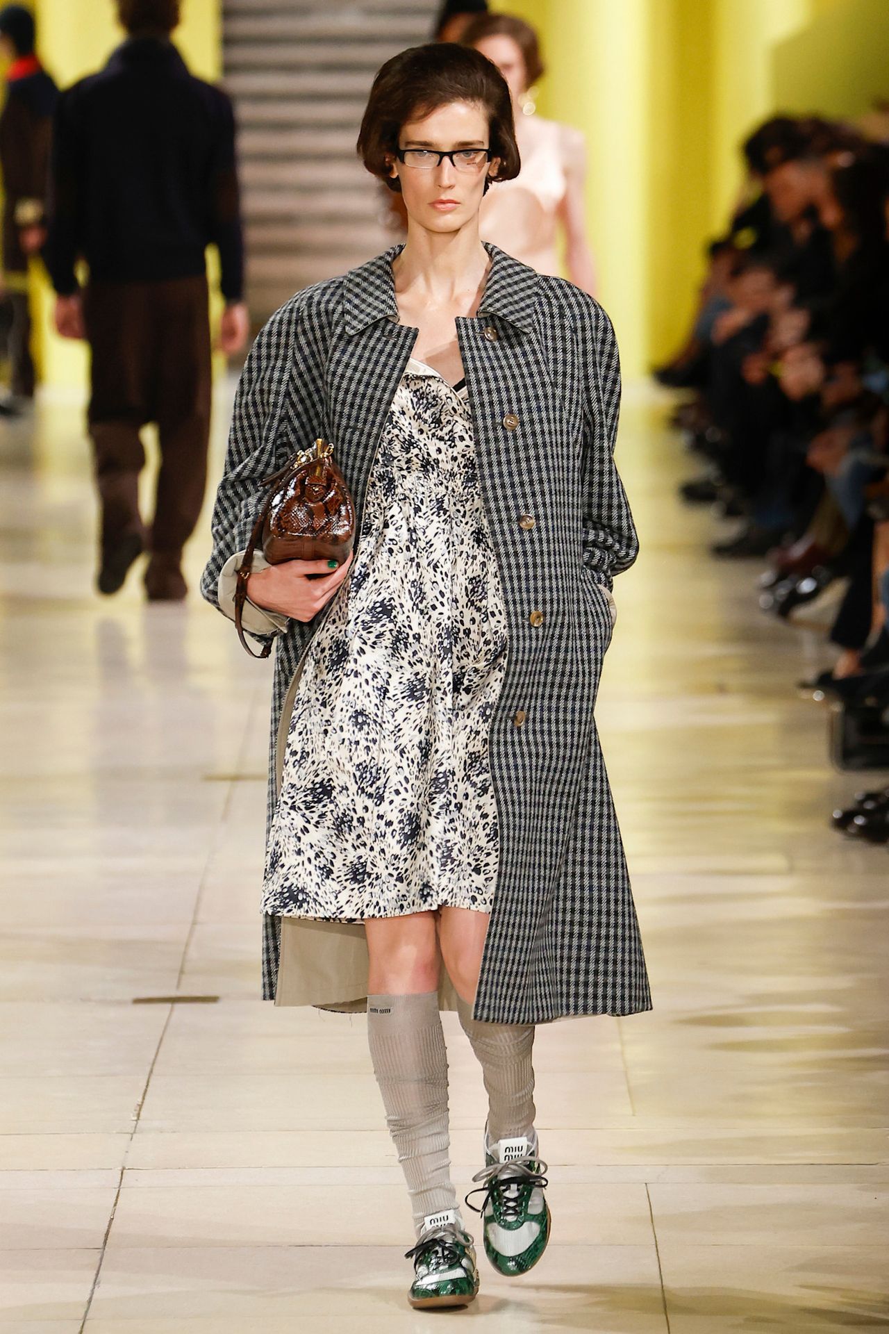 Model walking in the F/W 25 Miu Miu show during Paris Fashion Week wearing a plaid car coat with a floral dress.