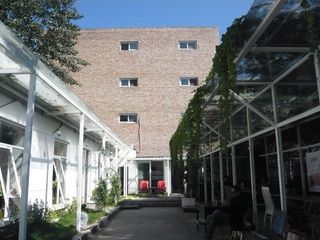 View between two low buildings with roofs overhanging with foliage, towards a larger brick building