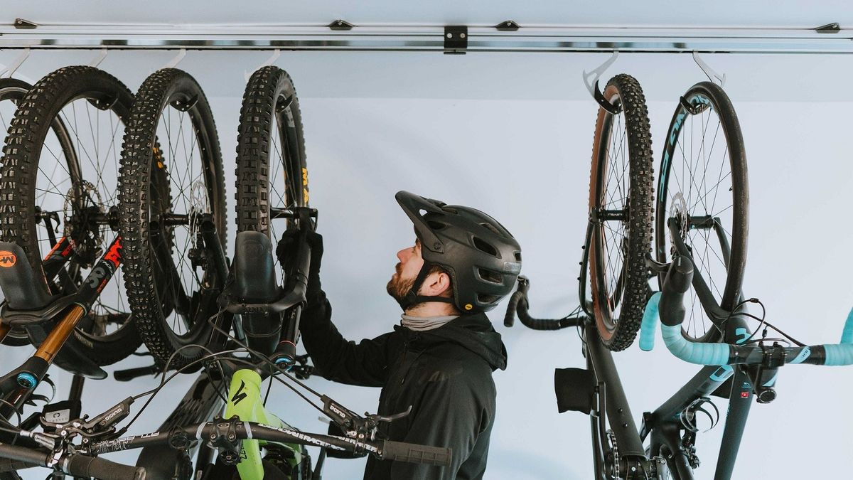 A man wearing a mountain biking helmet is standing between several mountain bikes hung up by their wheels, preparing to lift one off