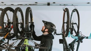 A man wearing a mountain biking helmet is standing between several mountain bikes hung up by their wheels, preparing to lift one off