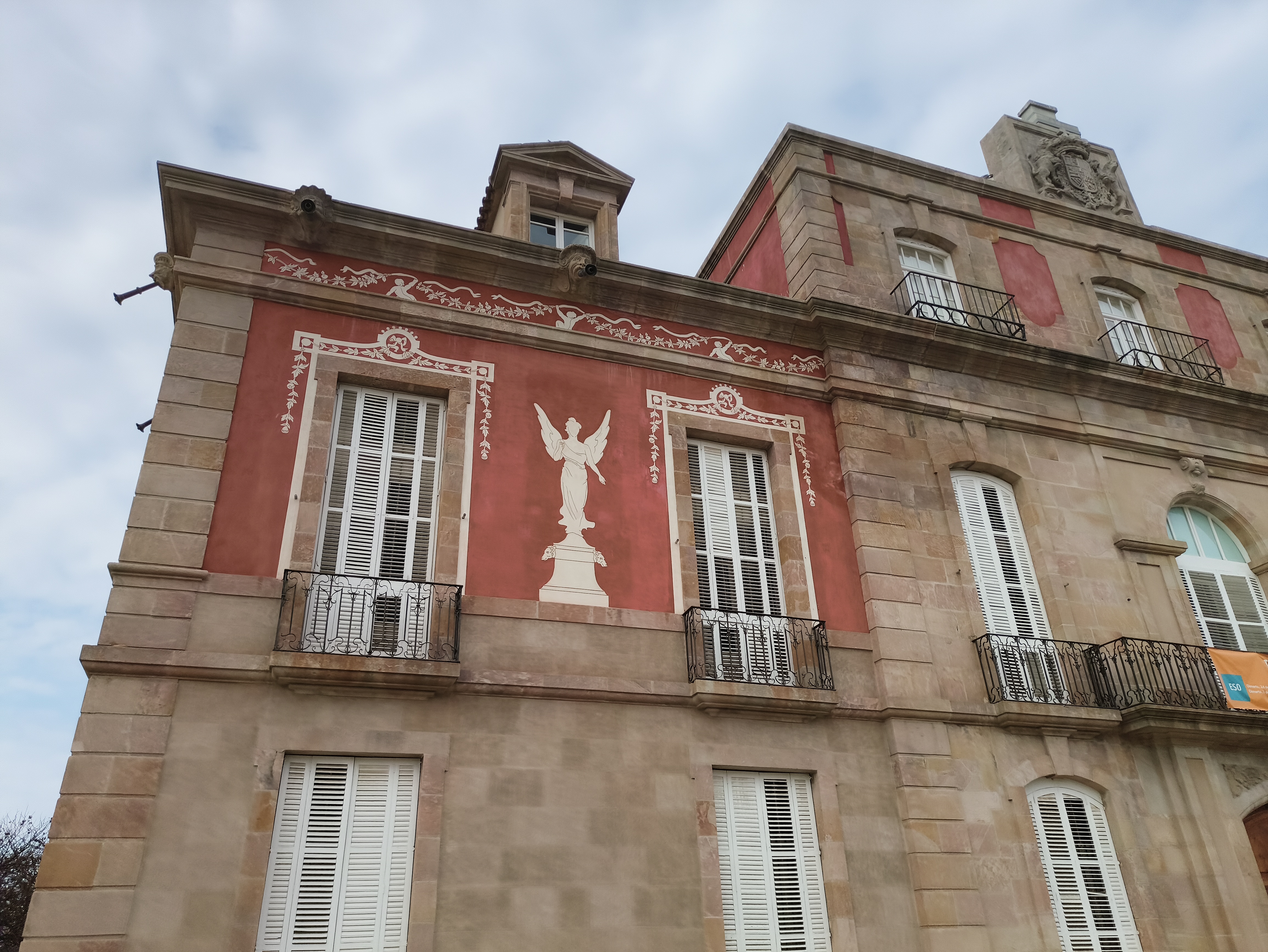The upper facade of a historic building features red panels decorated with white neoclassical figures and patterns, flanked by tall windows with white shutters and wrought-iron balconies.