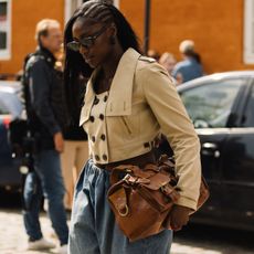 Woman wears cropped trench coat, blue trousers and carries brown Mulberry bag