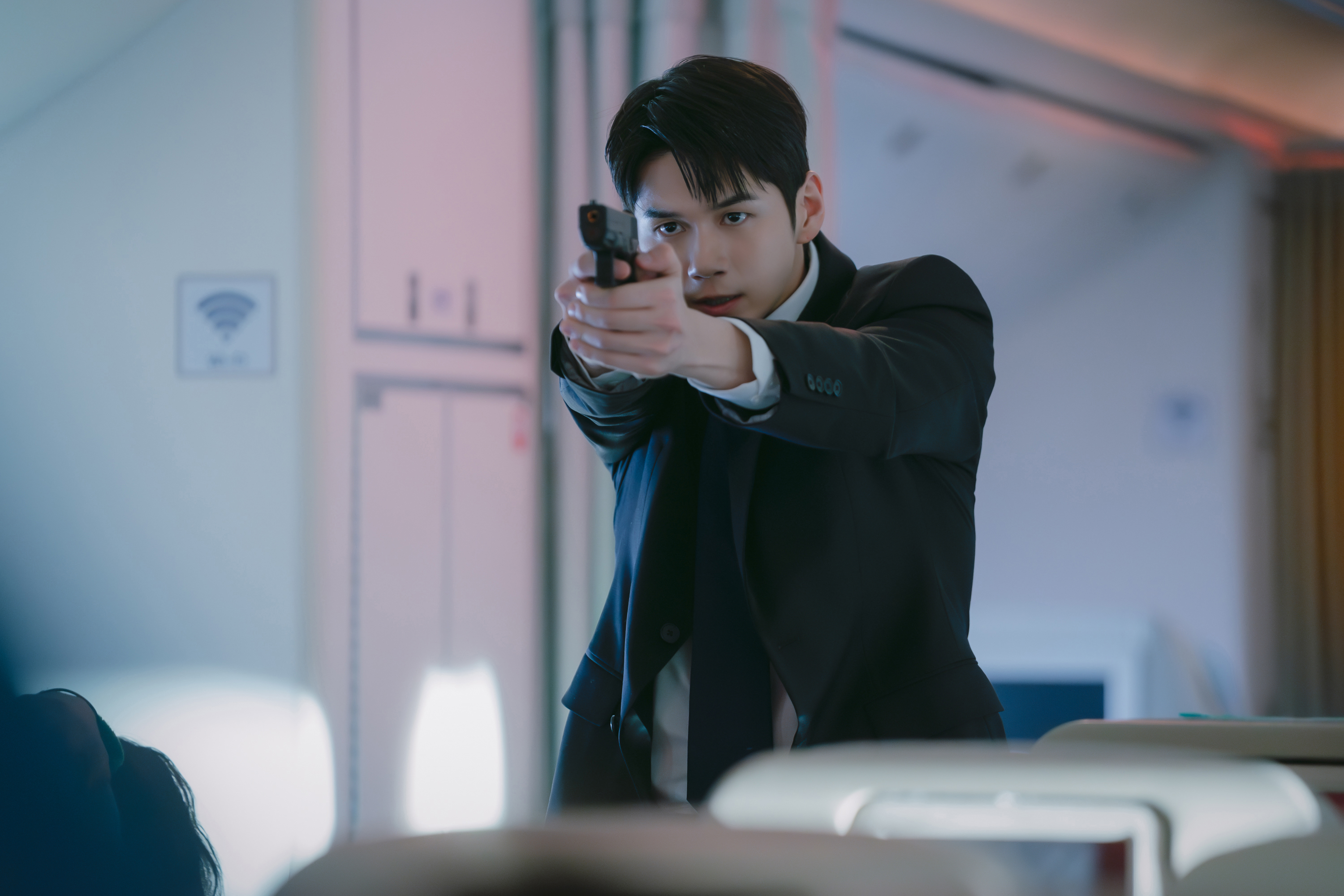 A man (Ong Seong-wu as Bae Hyeon-woo) aims a gun while standing in an airplane aisle, in the Netflix K-drama 'Boyfriend on Demand.'