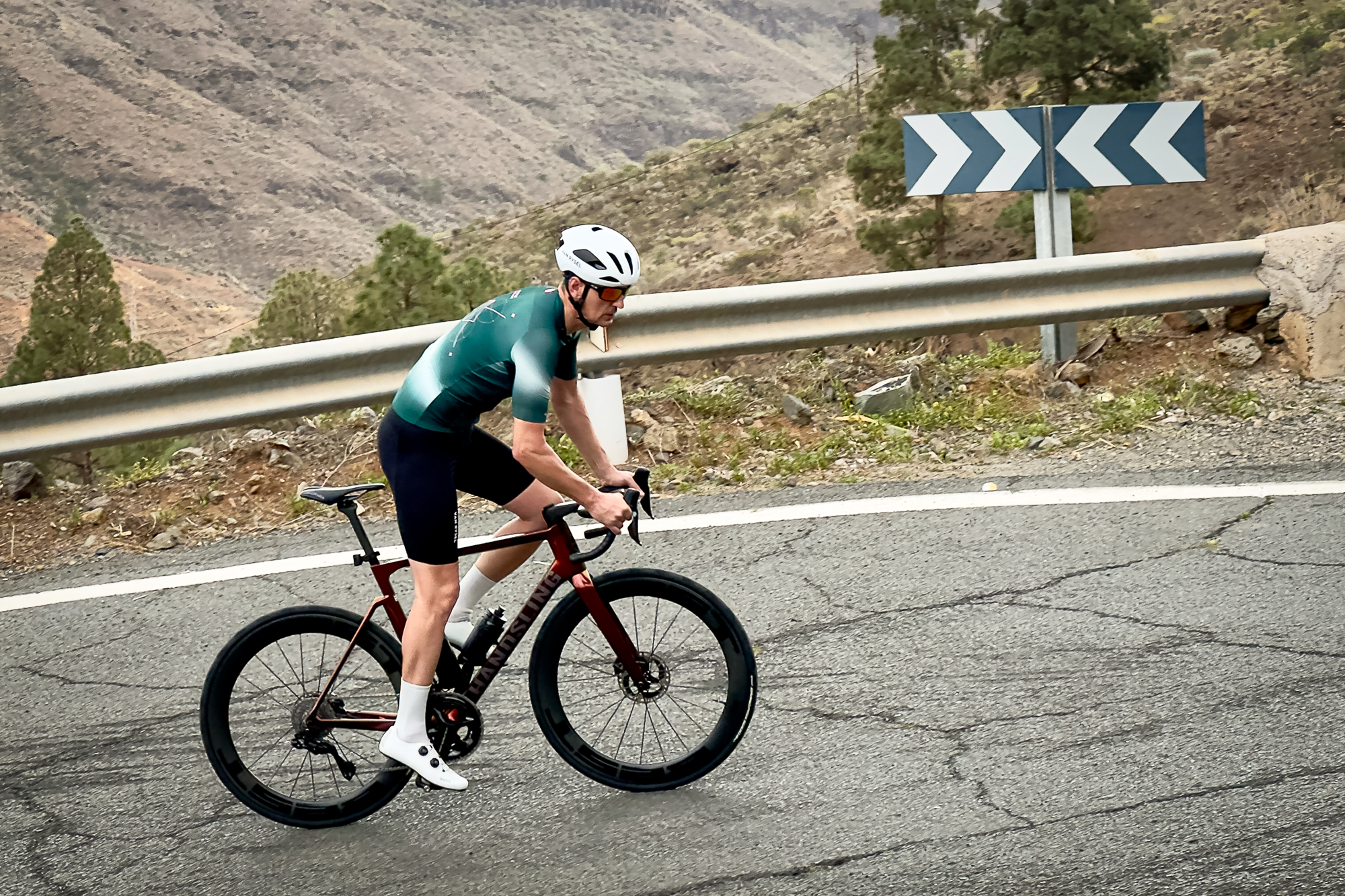 Man wearing a green jersey, black shorts and a white helmet riding an orange road bike up a hill out of the saddle