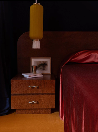 Image of a dark, moody bedroom. There is a bed with a wooden extended headboard with red, silk bedding. There is a matching wooden bedside table that has wavy, silver drawer pulls.