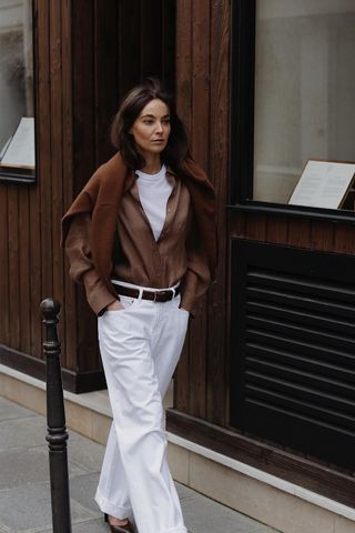 how to make a button-down shirt look more expensive is shown in a photo of a woman walking down the sidewalk in Paris wearing a brown cardigan layered over her shoulders styled with a white t-shirt under a brown button-down shirt, brown belt, white baggy jeans, and brown pumps