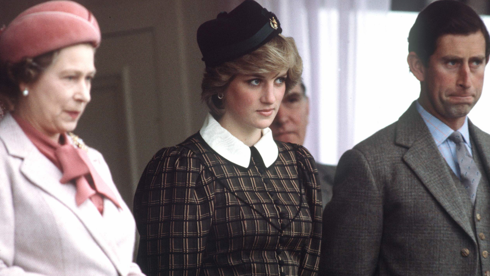 Princess Diana wears a tartan dress with a white collar and stands between Queen Elizabeth II, who wears a light pink coat, and King Charles