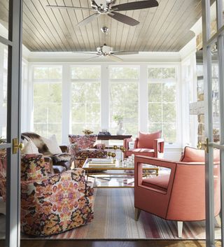sunroom with shiplap ceiling and fan, coral armchairs, floral armchairs, glass and brass coffee table, stripe rug, fireplace to right