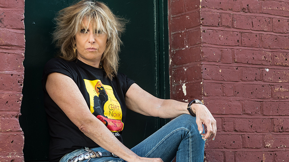 Close-up colour portrait of Chrissie Hynde sitting in a doorway in a red-brick wall