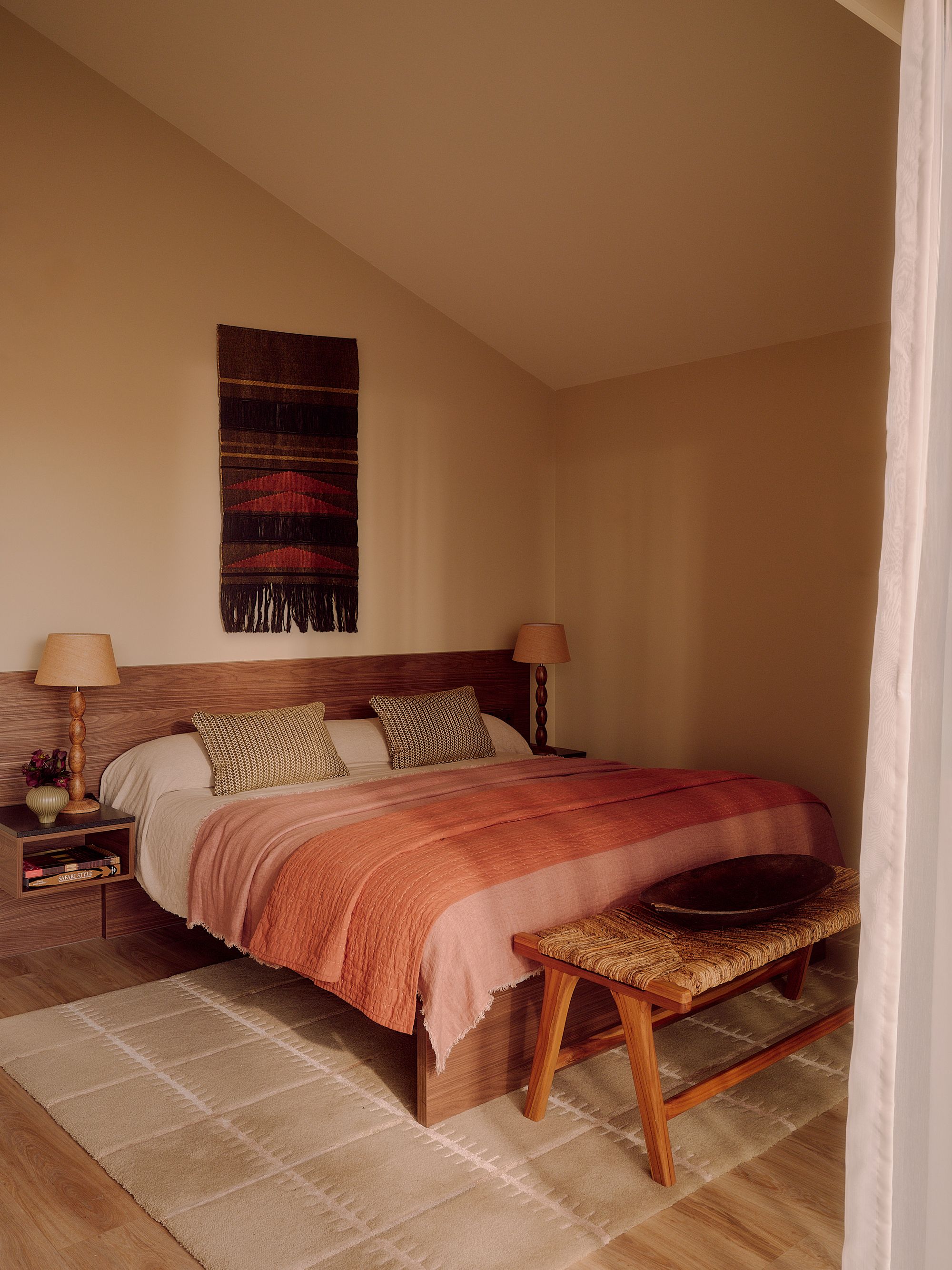 bedroom with neutral paint on the walls and ceiling, sloped roof, timber floors with beige rug, timber bedhead with built-in side tables styled with lamps, and timber and rattan bench at the end of the bed