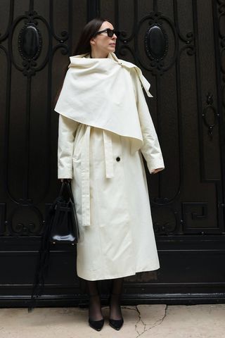 Spring trench coats shown in a photo of a woman standing in a doorway wearing black sunglasses with a cape trench coat, black dress, black sheer tights, black pumps, and a black structured handbag