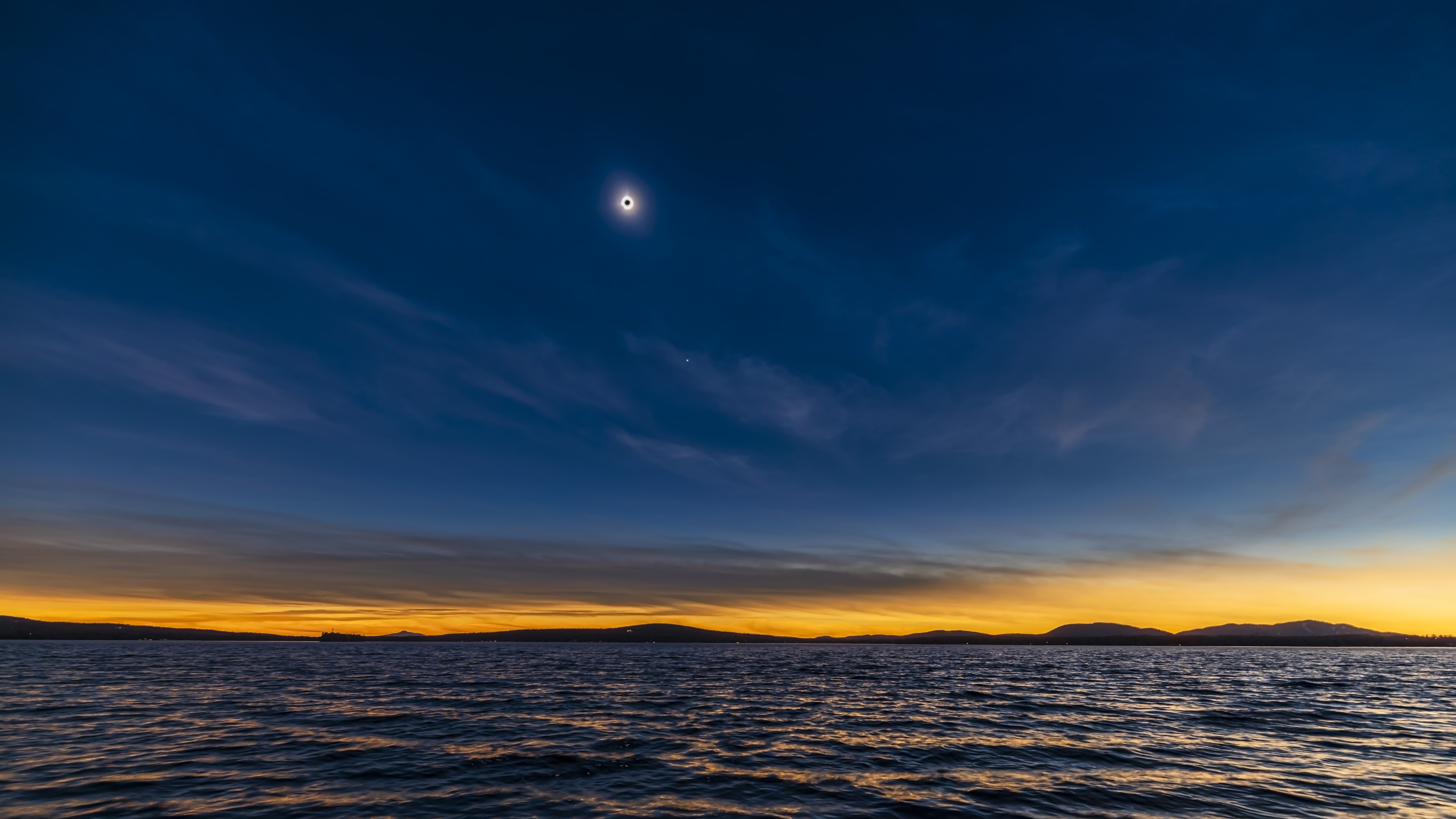 total solar eclipse over a large body of water and a orange sky near the horizon.