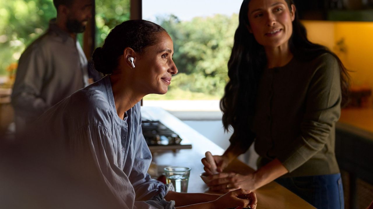 Woman wearing Apple AirPods Pro 2 as hearing aid, standing over kitchen island in house with friends