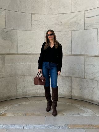 Nikki wears a black sweater, blue jeans, brown suede knee-high boots, and a brown bally bag.