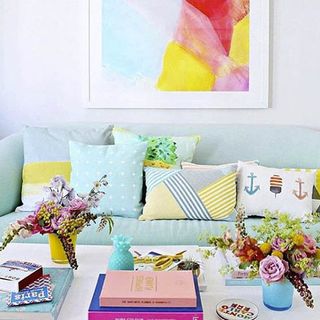 A baby blue sofa covered in colourful cushions sits behind a white table sporting colourful flower arrangements and books.