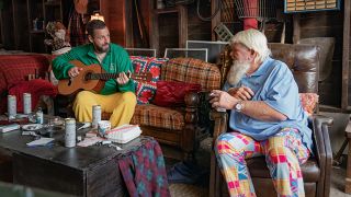 John Daly in polo and wild pants looking at Adam Sandler on a couch with his guitar in Happy Gilmore 2.