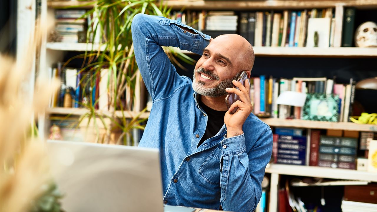 An older man smiles as he talks on the phone while sitting at his desk at home.