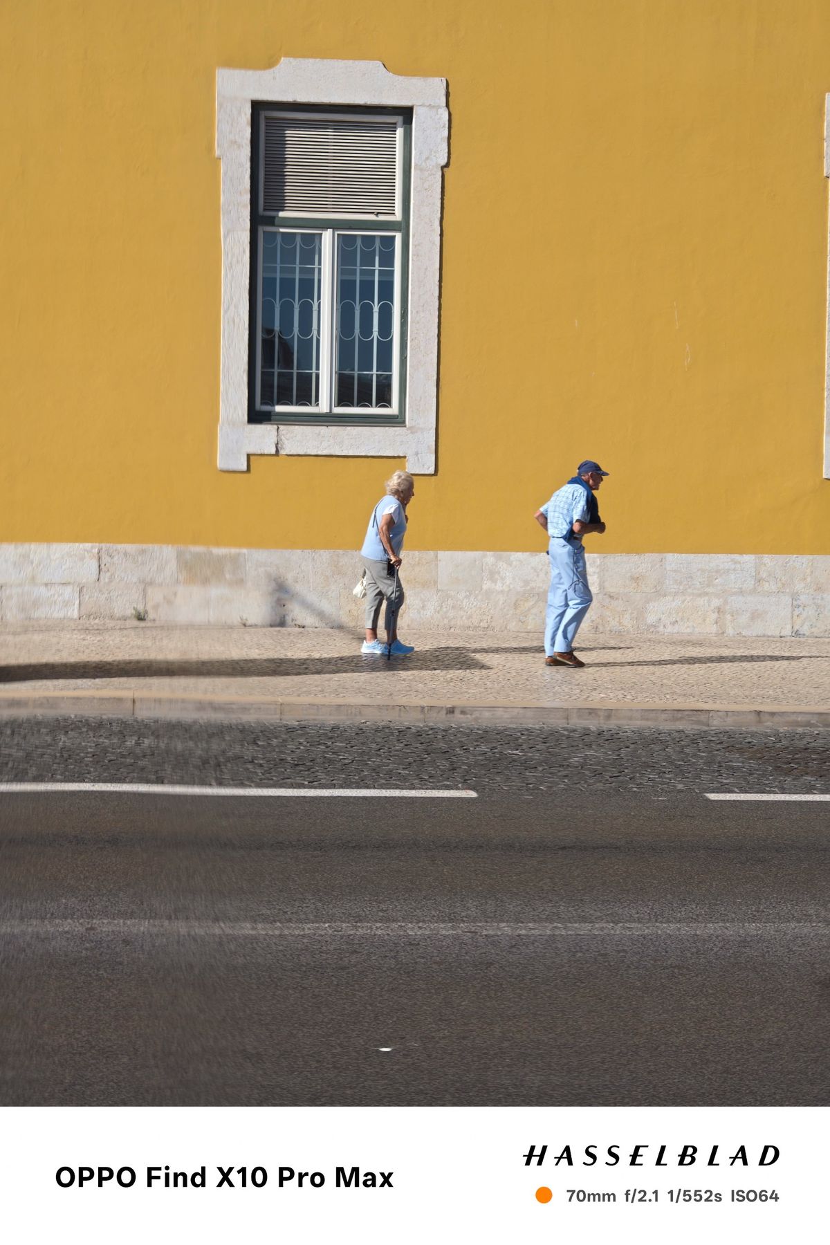 Elderly couple walking beside a bright yellow building in strong sunlight.
